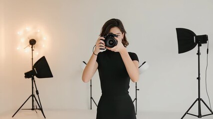 Young female photographer in black shirt holding a camera in a professional photo studio with lighting equipment in the background, symbolizing creative work and visual arts
