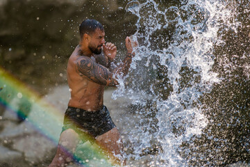 Fototapeta premium Powerful man on nature. Sexy shirtless man splashing under waterfall. Fit and strong male body in nature. Sexy model enjoying summer. Male beauty. Muscular torso with water drops. Topless guy in