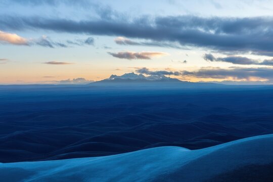 Serene mountain range emerges from vast blue rolling hills under dramatic clouds at dawn, evoking tranquility and adventure. - Powered by Adobe