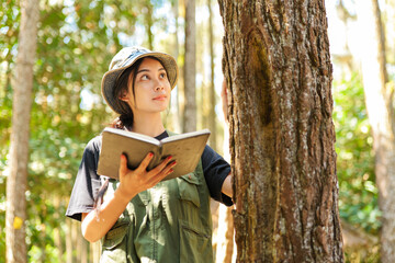 A woman is standing next to a tree and holding a book. She is wearing a green vest