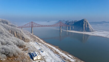 Fototapeta premium Winter panorama of a bridge over a river. Frozen landscape with a red-steel bridge, snow-covered banks, and misty mountains