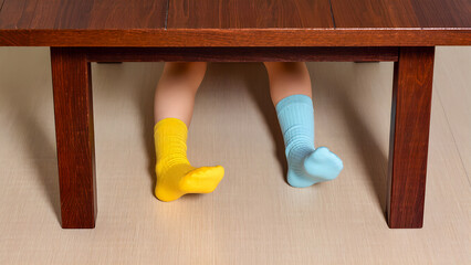A close-up of the feet of a person, presumably a child, peeking out from under a dark brown wooden table. The person is wearing multi-colored socks: one is bright yellow, the other is light blue. The 