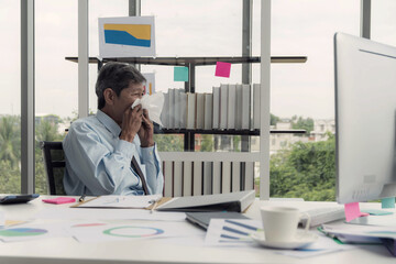 Mature man in a business shirt, clearly suffering from cold or allergies, uses a tissue at his...