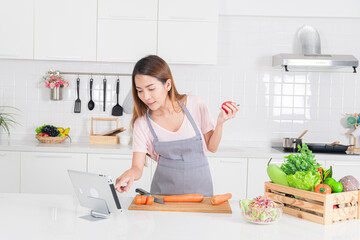 Attractive woman is using a digital tablet to follow a recipe while actively preparing fresh ingredient in like carrot and holding a tomato in a bright modern white kitchen.