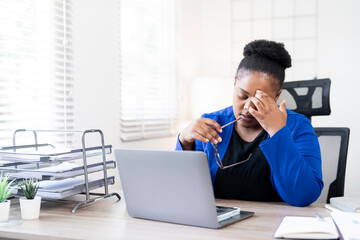 Stressed African American woman holding glasses and rubbing eyes while working on laptop in modern...