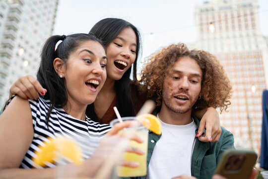 Excited friends watching something on a smartphone while drinking cocktails on a rooftop bar
