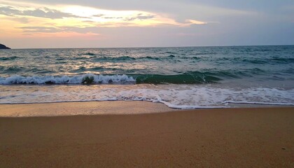 Obraz premium Gentle waves lap a sandy beach at sunset, with a darkened headland visible in the distance.