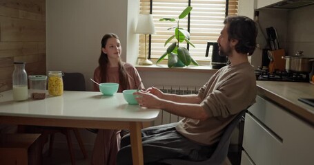 Side view of teenage girl sitting at table with her dad and chatting after morning breakfast with cereal and milk in cozy kitchen in the morning - Powered by Adobe