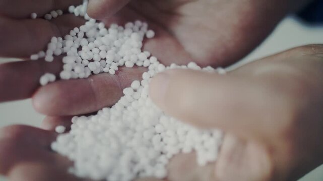 An extreme close-up shot of a person's hands inspecting white, spherical fertilizer granules. The pellets are poured from one hand to another, some falling in slow motion