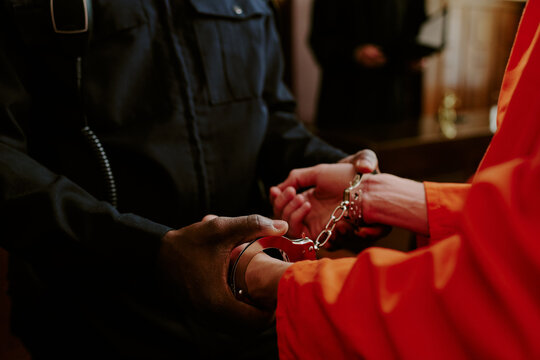 Black male police officer handcuffing Caucasian male prisoner in orange jumpsuit during arrest process, both standing indoors with focus on hands and handcuffs, law enforcement concept