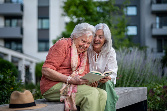 Elderly mother sitting on bench with adult daughter, reading book.