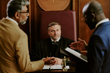 Middle aged Caucasian male judge sitting at bench listening to arguments from middle aged biracial man and Black man, standing in courtroom discussing legal case