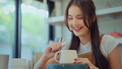 Asian woman painting ceramic art and pottery designs in her studio – happy artisan potter sitting with handmade tools, enjoying sculpting process and creative small business lifestyle