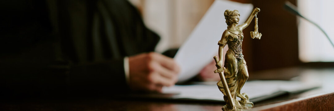 Caucasian middle aged man holding legal documents in courtroom background, bronze Lady Justice statue standing in foreground symbolizing law and justice, judge working at desk - Powered by Adobe