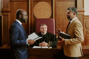 Caucasian middle aged male judge sitting at bench, observing Black middle aged man in suit holding...