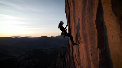 Climber at Sunset - A female rock climber ascends a steep cliff face during a stunning sunset. The vast mountain range provides a dramatic backdrop to this adventurous scene.
