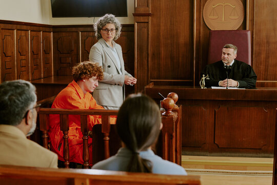 Caucasian teenage boy sitting in defendant box wearing orange jumpsuit, middle aged Caucasian woman standing and speaking, middle aged Caucasian male judge listening in courtroom