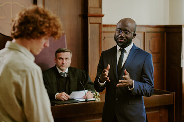 Black middle aged man in suit gesturing while speaking in courtroom, Caucasian teenage boy standing in foreground, Caucasian middle aged male judge sitting behind bench observing