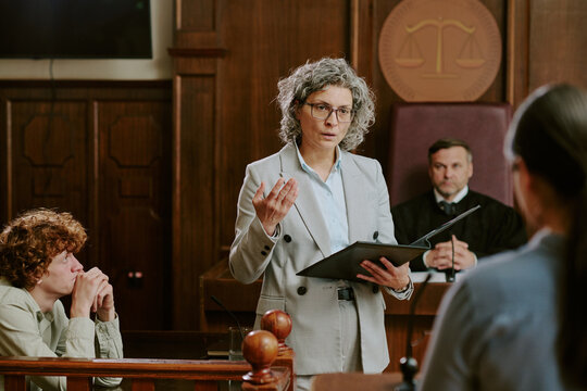 Fototapeta Caucasian middle aged woman lawyer addressing courtroom while holding folder, Caucasian teenage boy sitting at witness stand, judge and another woman present in background