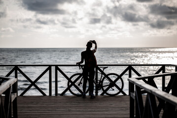 A woman stands on a wooden boardwalk by the sea, holding a smartphone up for a photo. 