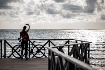 A woman stands on a wooden boardwalk by the sea, holding a smartphone up for a photo. 