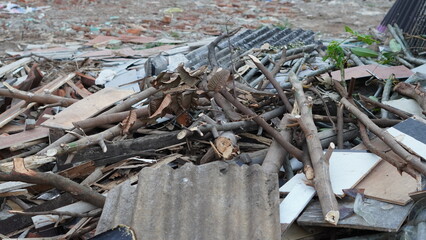 A pile of wooden branches, leaves, and construction debris including tiles and boards, discarded outdoors in an open area.