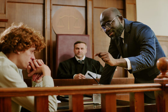 Black middle aged man in suit gesturing while addressing Caucasian teenage boy sitting at witness stand in courtroom with judge observing proceedings in background