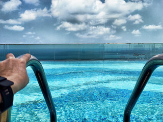 Hand reaching for pool ladder at rooftop infinity pool, capturing vibrant blue water and cloudy sky...