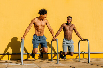 Two Latino men perform strength exercises outdoors against a vibrant yellow wall during a sunny day