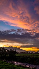 Obraz premium Vibrant sunset hues paint the sky above a tranquil landscape, with clouds reflecting warm colors over a field and stream