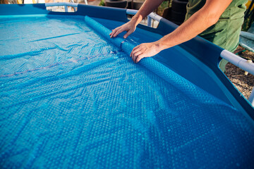 The blue pool cover is assembled into a roll by men's hands, the concept of summer recreation and...
