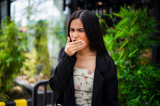 An inquisitive young asian woman breathing into her palm and smelling it, opening her mouth slightly while checking her breath at a cafe