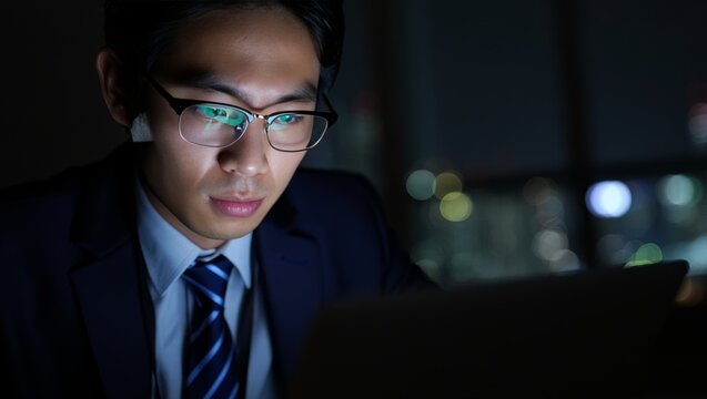 Asian businessman working late at night on laptop in office with city skyline view