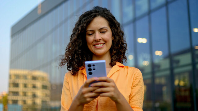 Smiling businesswoman using mobile phone app for online shopping or checking social media in front of modern office building