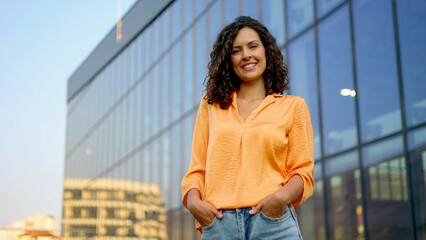 Fototapeta premium Professional businesswoman posing confidently near glass skyscraper, wearing tailored suit, hands in pockets, warmly smiling