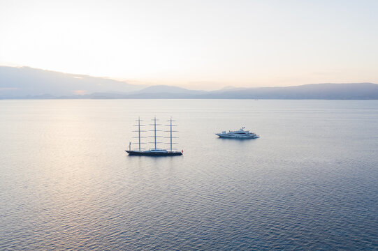 Idra, Greece - 30 July 2025: Aerial view of two majestic yachts cutting through the tranquil, deep blue waters under the soft glow of a serene dawn.