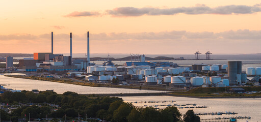 Fototapeta premium Industrial area in Copenhagen, Denmark, at sunset with chimneys, storage tanks, wind turbines, a waterway with boats, greenery, and distant cranes.
