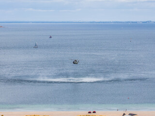A military helicopter hovers low over calm blue water near Copenhagen, creating ripples. A sandy...