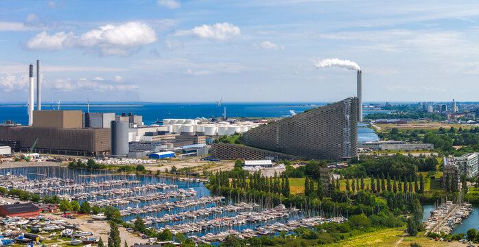 Aerial view of Copenhagen, Denmark, showing CopenHill waste to energy plant, a marina with boats, wind turbines, industrial areas, and the sea.