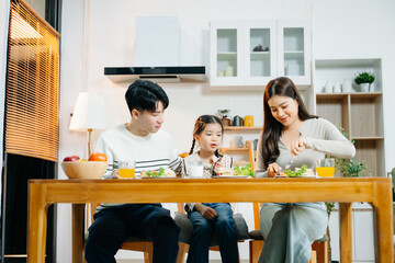 Asian family shares loving breakfast time. Parents help daughter drink milk while bonding over food and smartphone in a cozy kitchen.