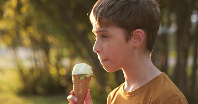 Child boy eating ice cream in park at sunset. Happy child enjoys ice cream outdoors during golden hour. Child with ice cream smiles in nature, enjoying peaceful summer evening. Colorful ice cream cone