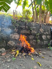 Bright orange flames burning a pile of dry leaves near a stone wall in an outdoor rural area.