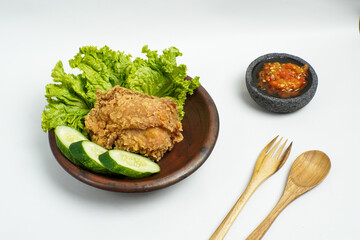 Crispy fried chicken served with fresh lettuce, cucumber, and spicy chili sambal in a stone mortar, traditional Indonesian food on a clay plate.