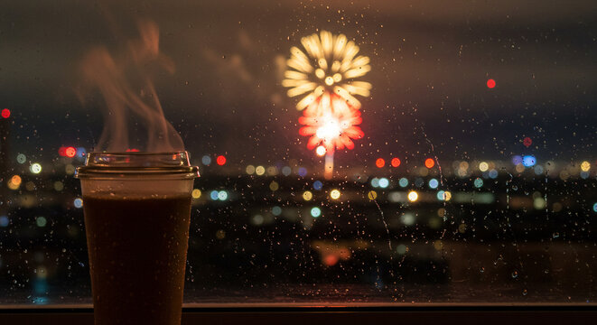 A steaming cup of coffee sits on a windowsill as fireworks explode over a city skyline at night, seen through a rain-streaked window.