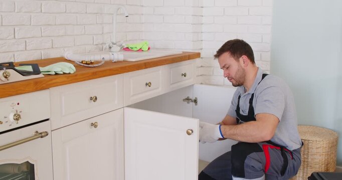 Plumber working in kitchen, trying to repair sink or solve problem with leaking, repairing pumps, dressed in overalls, working in service industry, examining all details. Plumbing concept