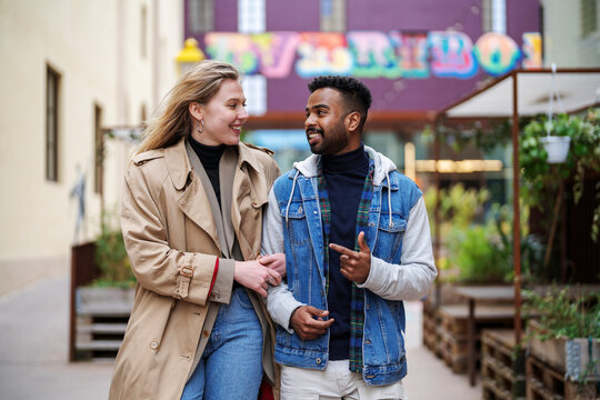 Happy multiracial couple wearing casual clothes and outerwear talking to each other while strolling close during date in city