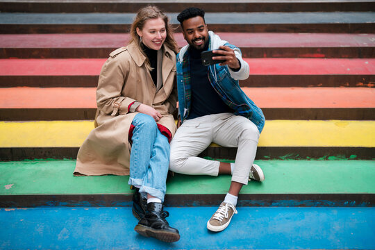 Full body of multiracial cheerful couple sitting on colorful staircase painted in colors of LGBT flag and taking self portrait on cellphone during date