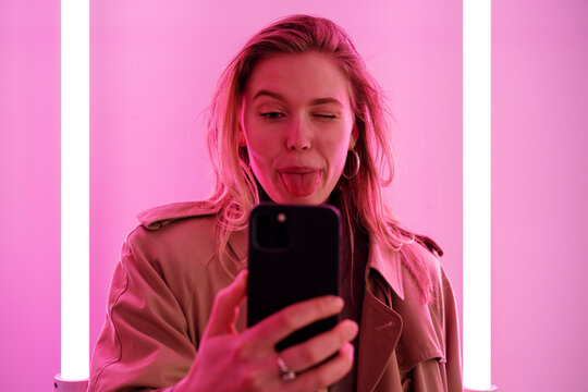 Playful female winking and showing tongue while taking self portrait on cellphone against wall with bright pink neon lamps