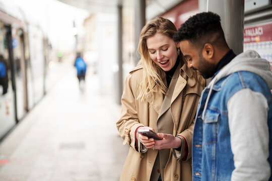 Young woman in trench coat using cellphone and showing to Arab boyfriend while standing together on public transport stop during date