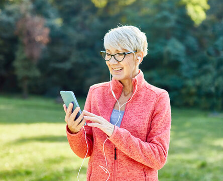 Happy active senior woman having fun using smartphone and wearing earphones and sportswear, checking music or an exercise app, after having an exercise sport activity outdoors